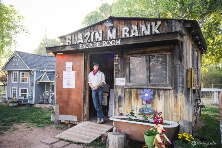 Man standing outside rustic escape room, 'Blazin' M Bank', with wooden facade and decorative bathtub.